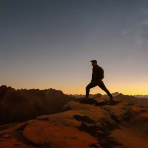 An image depicting a solitary adventurer standing atop a rugged mountain peak at dusk, surrounded by an expansive wilderness